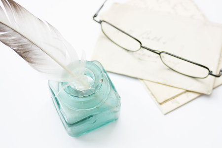 A feathered quill pen standing in a glass ink pot with old hand written letters behind with a pair of black framed reading glasses set on a white backgroundの写真素材