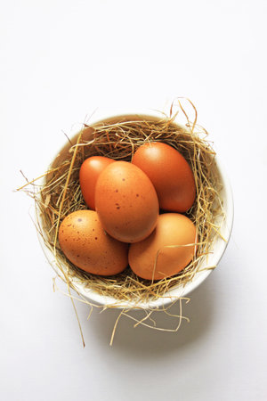 Five freshly laid organic brown eggs, set in a white ceramic bowl with straw. Set on an isolated...