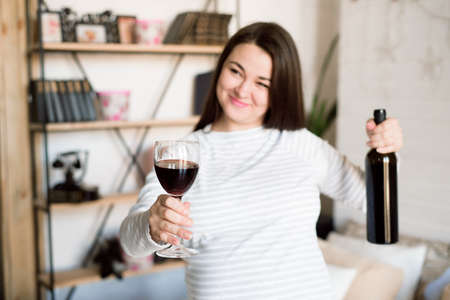 Pregnant alcoholic woman is pouring wine from a bottle into a glass and drinking.の写真素材