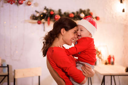 Happy mother and adorable baby in Santa hat celebrate Christmas. New Years holidays. Toddler with mom in the festively decorated roomの写真素材