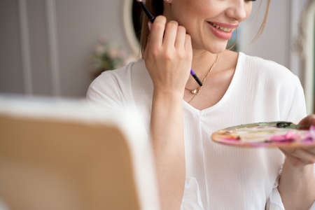 Painter drawing in art studio using easel. Portrait of a young woman painting with oil paints on white canvas, side view portraitの写真素材