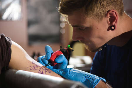 Master tattoo draws the black paint on the clients tattoo. Tattoo artist holding a tattoo machine in blue sterile gloves and working on the professional matの写真素材