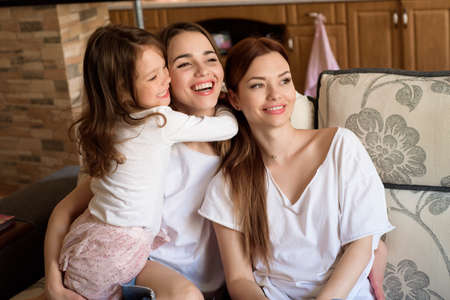 Portrait of two sisters and a little girl sitting on the couch, smiling. Concept: family, sisters, home and happiness.の写真素材