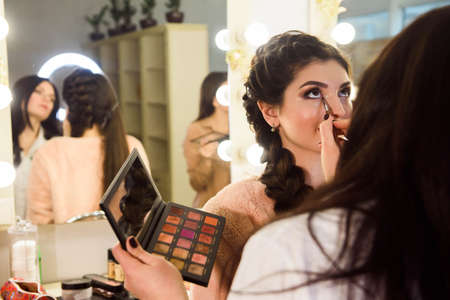 Make-up artist applying bright base color eyeshadow on models eye and holding a shell with eyeshadow...