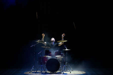 Drummer in a cap and headphones plays drums at a concert under white light in a smoke.