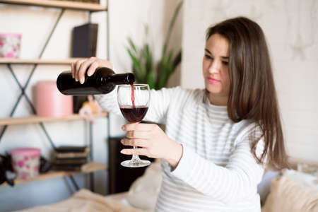 Pregnant alcoholic woman is pouring wine from a bottle into a glass and drinking.の写真素材