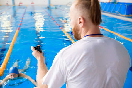 A young swimming coach with a stopwatch in his hand watches the training session near the sports pool.の写真素材