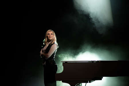 Portrait of young caucasian woman standing near the grand piano in the beam of light