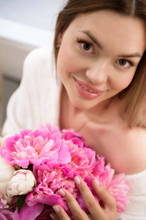 Beautiful girl in a white bathrobe with a bouquet of pink peonies near the bath.の写真素材