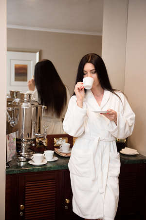 A girl in a white bathrobe is resting and drinking a cup of coffee or tea enjoying wellness weekendの写真素材