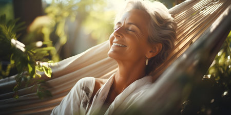 Happy senior woman enjoying technology in hammock surrounded by garden sunlight. Concept Outdoor Photoshoot, Technology, Seniors, Hammock, Gardenの素材