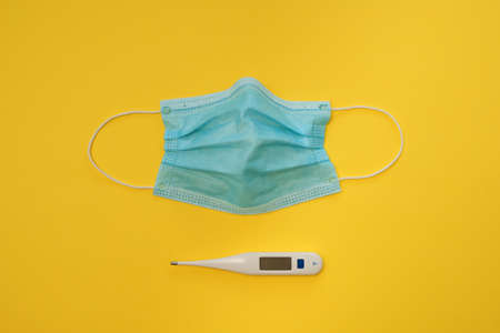 COVID-19 disposable surgical face mask and digital thermometer on yellow background. Protection against coronavirus and epidemic. Medical conceptの写真素材