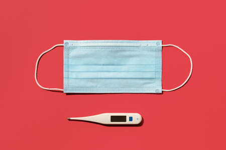 COVID-19 disposable surgical face mask and digital thermometer on red background. Protection against...