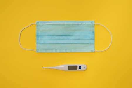 COVID-19 disposable surgical face mask and digital thermometer on yellow background. Protection against coronavirus and epidemic. Medical conceptの写真素材