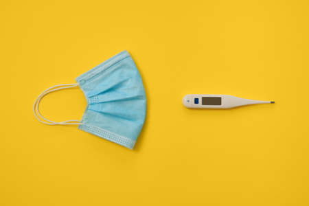 COVID-19 disposable surgical face mask and digital thermometer on yellow background. Protection against coronavirus and epidemic. Medical conceptの写真素材