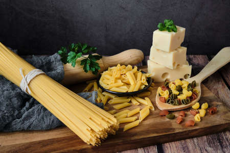 Different raw uncooked Italian pasta and cheese on wooden table against dark background. Diet and food concept.の写真素材