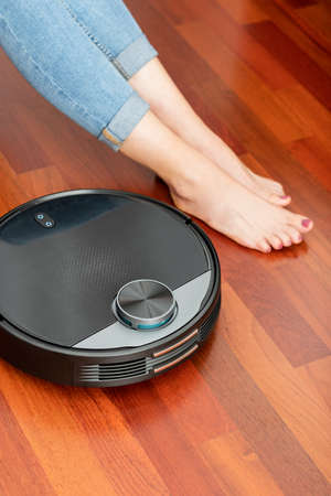 Young girl feet when a round smart automatic vacuum cleaner passes to clean the floor, controlling machine housework robotの写真素材