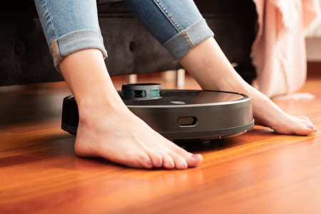 Young girl feet when a round smart automatic vacuum cleaner passes to clean the floor, controlling...