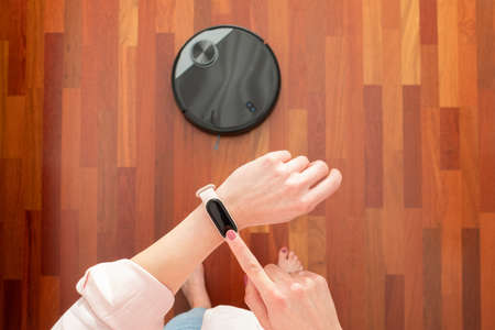 Top view of faceless middle section of young woman using smart watch to control an automatic vacuum cleaner to clean the floor, machine housework robotの写真素材