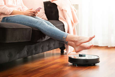 Faceless middle section of young woman using automatic vacuum cleaner to clean the floor, controlling smart machine housework robotの写真素材