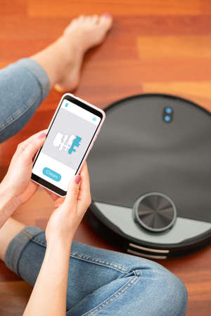 Young girl feet at home using a smart phone to control a round smart automatic vacuum, machine housework robot cleaning the floorの写真素材