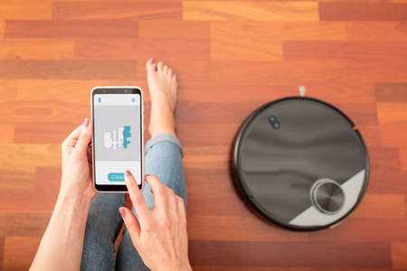 Young girl feet at home using a smart phone to control a round smart automatic vacuum, machine housework robot cleaning the floorの写真素材