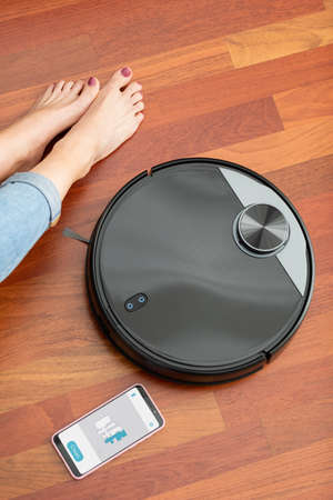 Faceless middle section of young woman with smartphone and automatic vacuum cleaner at the floor. Housework robot with app mockupの写真素材