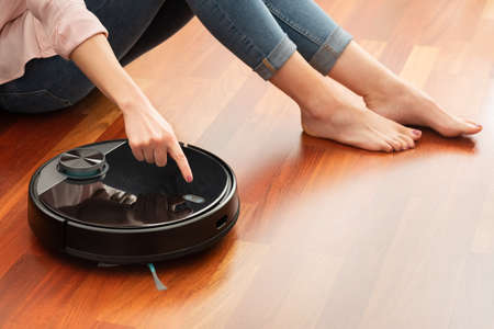 Faceless middle section of young woman using automatic vacuum cleaner to clean the floor, controlling smart machine housework robotの写真素材