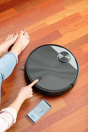 Faceless middle section of young woman touching an automatic vacuum cleaner to clean the floor, controlling smart machine housework robot with smartphone appの写真素材