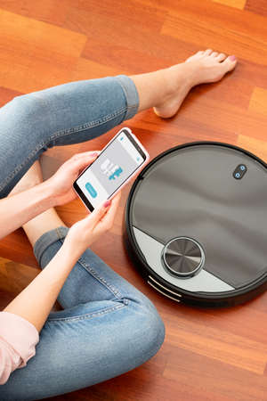 Young girl feet at home using a smart phone to control a round smart automatic vacuum, machine housework robot cleaning the floorの写真素材