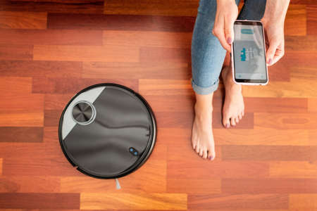 Young girl feet at home using a smart phone to control a round smart automatic vacuum, machine housework robot cleaning the floorの写真素材