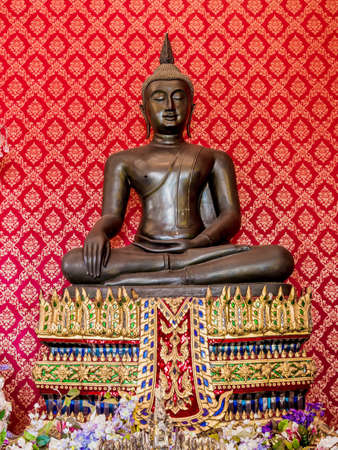 Bronze Buddha statue with red wall behind in the temple.のeditorial素材