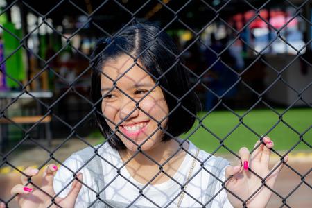 Asian young girl behind chain link fence looking for somone or seeing sport gameの写真素材