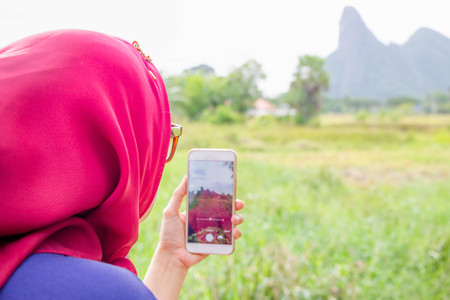 Muslim woman in red scarf (Hijab) look from back taking  photo of natural scene by smartphoneの写真素材
