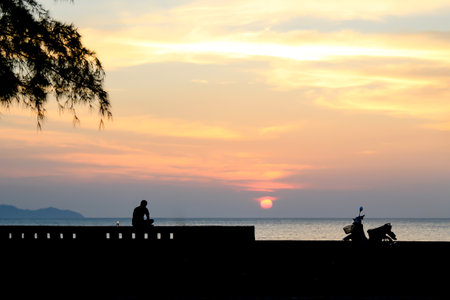 man sitting alone on the beach drinking and watching sunset with his motorcycleの写真素材