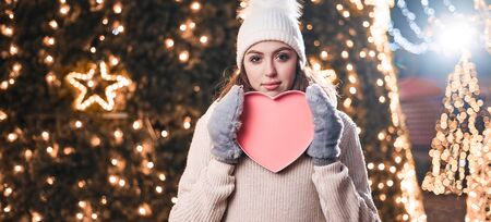 Portrait of pretty girl holding heart. She in knitted hat, warm sweater among the night lights. She holds red heart in gloves, smiling. Concept for 14 February.の写真素材