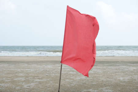 Selective focus picture of waving red flag at the beach with strong water wave insight. In Malaysia,...