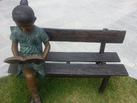 A statue of a girl reading a book while sitting on the wooden bench with muffler around neck on a sunny day.の写真素材