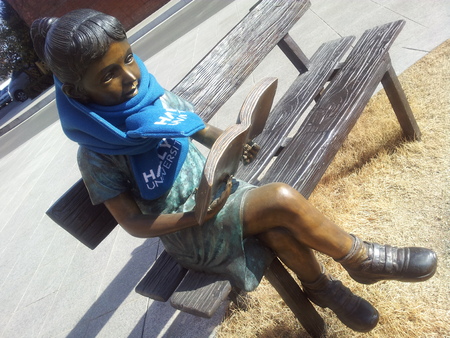A statue of a girl reading a book while sitting on the wooden bench with muffler around neck on a sunny day.の写真素材