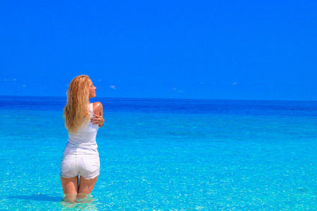 Gorgeous blond woman with white clothes is standing in the water at the sandy beachの写真素材