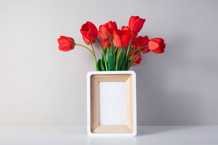 White mockup photo frame on a white table and a wall. A vase of green flowers next to the frame....