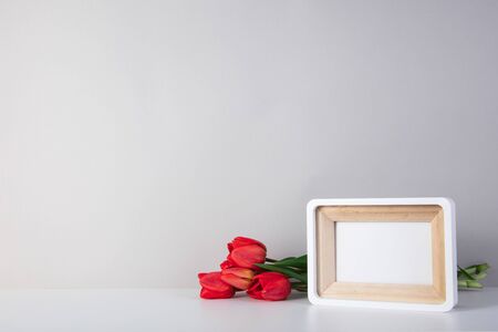 White photo frame on a white table with red tulips. Spring with copy space.