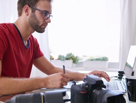 Handsome young man with a beard sitting at his glass desk in his home office busy editing images on his laptop with his camera visible in the foreground.の写真素材