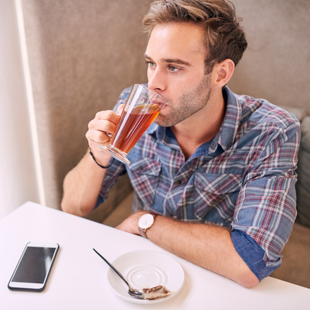 handsome young man taking a sip of his tea while sitting next to the window in a modern well lit cafe while wearing a check shirt, with his phone on the table infront of himの写真素材