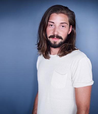 Young white man with long loose brown hair and a beard standing in front of a blue background while...