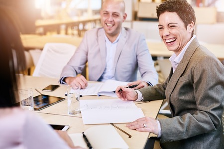 over the shoulder image of business executives laughing happily during a meeting seated at a conference table looking toward the camera.の写真素材