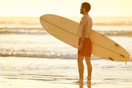 Young caucasian male wearing red shorts standing in the shallow waves on the beach while holding a surfboard perpendicular to himself.の写真素材