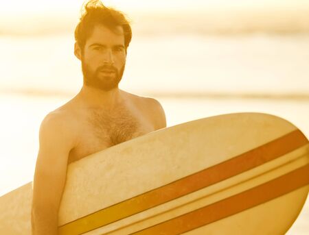 Young bearded caucasian male looking at the camera while holding a retro surfboard under his arm with the ocean waves in the background behind him.の写真素材