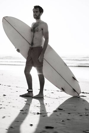 Black and white image of young male surfer standing on beach with his surfboard under his arm and...