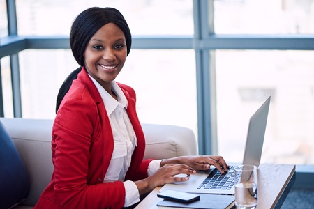 Black female businesswoman smiling at the camera while seated on a couch in a business lounge with her hands on her laptop keyboard with large windows and the cityscape behind her.の写真素材